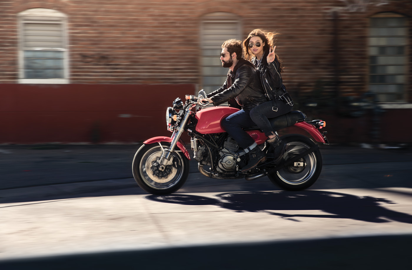 A man drives a red motorcycle while a woman sits behind him and throws up a peace sign.