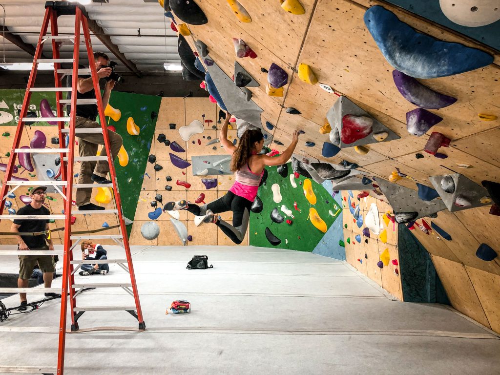 Olympic climber Brooke Raboutou hangs off a hold on a bouldering wall while photographer Tyler Stableford stands on a ladder.