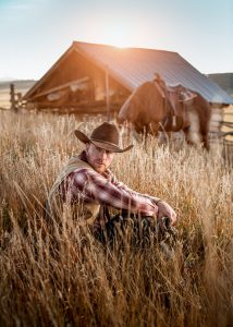This Dramatic Image of a Modern Cowboy at Sunset is a Picture of Life in the West. His Horse Grazes Quietly Near a Barn in the Tall Grass.