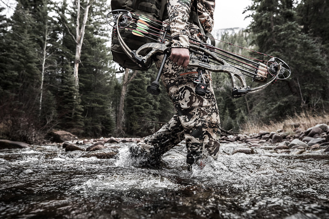 A big game bow hunter crosses a mountain stream while hiking in the alpine hunting elk.