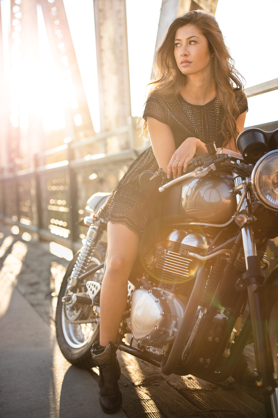 Woman sits on motorcycle, leaning forward and looking off-camera with bright light in background.