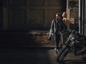 A man and woman lean against brick wall with motorcycle in foreground.