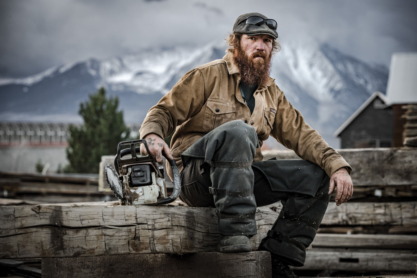 Man with long beard sits on wood wearing Patagonia workwear, with mountains in the background.