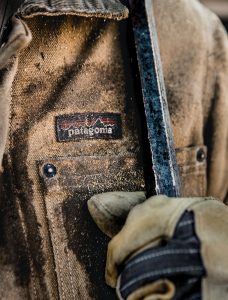 A gloved hand holds a metal bar up to the chest of a tan Patagonia workwear jacket.