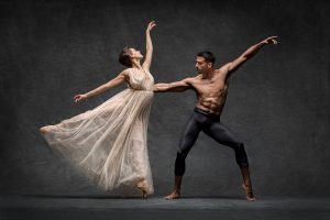 Two ballet dancers strike poses while dancing against dark backdrop.