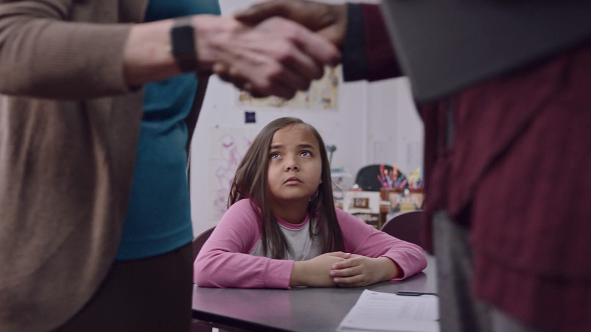 Girl looking up at a handshake between 2 adults, looking sad.