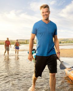 Josh Duhamel smiles next to a kayak, standing in a river with a paddle in hand.