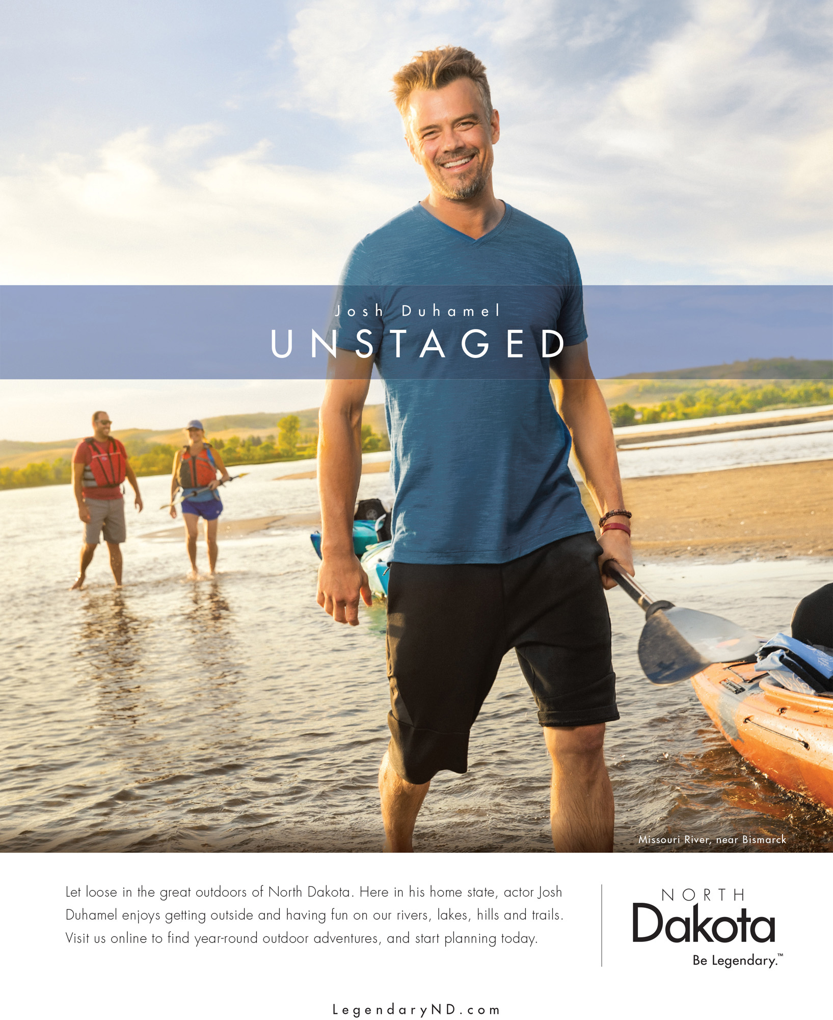 Josh Duhamel smiles next to a kayak, standing in a river with a paddle in hand.